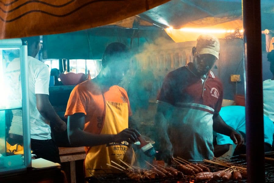Deux grilleurs préparent brochettes et saucisses dans la fumée, sous la bâche d’un maquis
